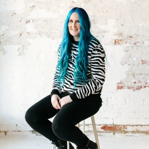 Bridget with blue hair sitting on a stool, with a white brick background.